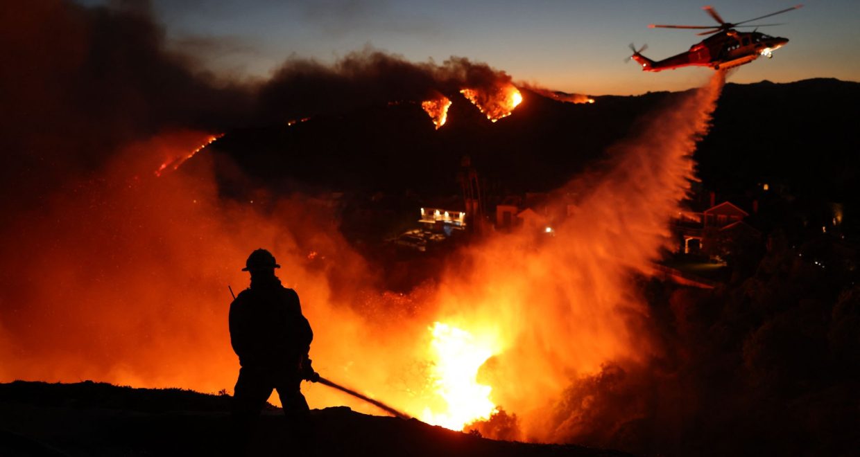 A firefighter is silhouetted in the glowing orange and yellow blaze of a wildfire as a helicopter above dumps a stream of water below — Image credit: California's wildfire season is shifting, with more blazes after the traditional high-risk window, study finds / https://www.livescience.com/author/stephanie-pappas